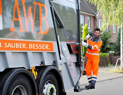 Auf der Triffläche eines Müllautos mit AWB Schriftzug steht ein Müllwerker und lächelt mit Daumen hoch in die Kamera. 